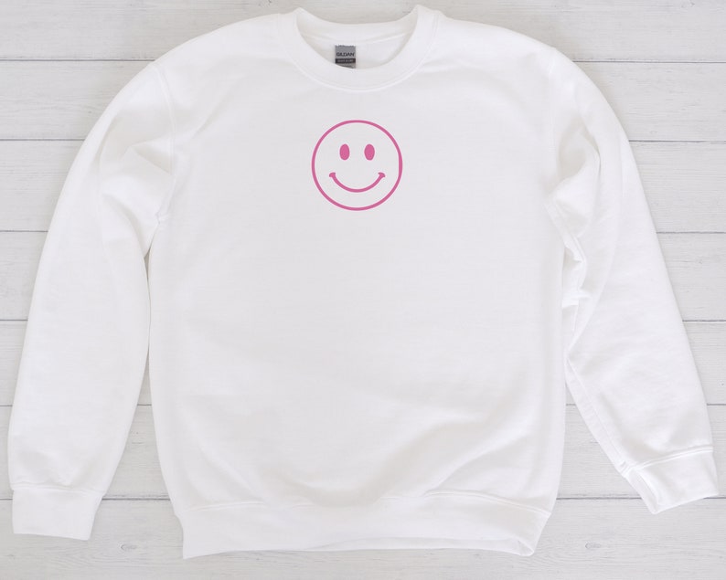 May include: White sweatshirt with a pink smiley face graphic on the front.