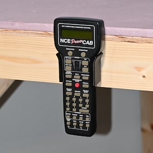 May include: A black NCE PowerCAB DCC compatible command control handheld throttle with a digital display screen, a central red emergency stop button, a speed slider, and numerous labeled grey buttons for model train locomotive operation.