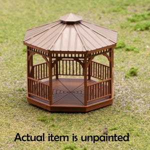May include: A miniature brown wooden gazebo with a sloped roof and decorative railings. The gazebo is unpainted and sits on a grassy surface. The text "Actual item is unpainted" is at the bottom.
