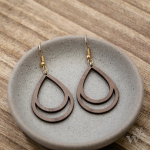 May include: A pair of wooden teardrop earrings with a double layer design. The earrings are made of dark brown wood and have gold-toned hooks.
