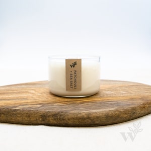 May include: A clear glass candle jar with a white candle inside. The jar has a brown paper label that says "Patchouli + Sea Salt". The candle is sitting on a wooden cutting board.