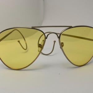 May include: Aviator-style sunglasses with a gold-colored metal frame and yellow-tinted lenses. The lenses are teardrop-shaped. The bridge and temples are thin, and the frame has a double bar above the nose bridge.