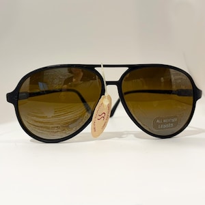 May include: Black aviator sunglasses featuring a double bridge design and brown-tinted lenses. A small, oval-shaped tag with the text SS is attached to the bridge. A sticker on the right lens reads ALL WEATHER LENSES in white text on a dark background.