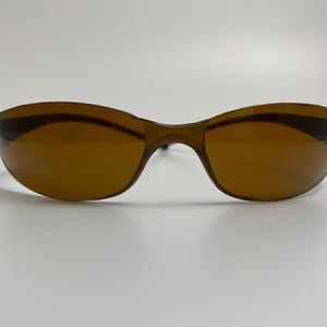 May include: Brown-tinted sunglasses with a curved, wraparound design. The lenses are a warm brown color, and the frames are a darker shade of brown. The sunglasses are set against a plain white background.