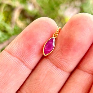 Peut inclure: Un petit pendentif doré avec une pierre précieuse rose foncé de forme marquise. Le pendentif est suspendu à une petite boucle. La pierre précieuse est sertie dans une lunette dorée. L'arrière-plan est un vert flou.