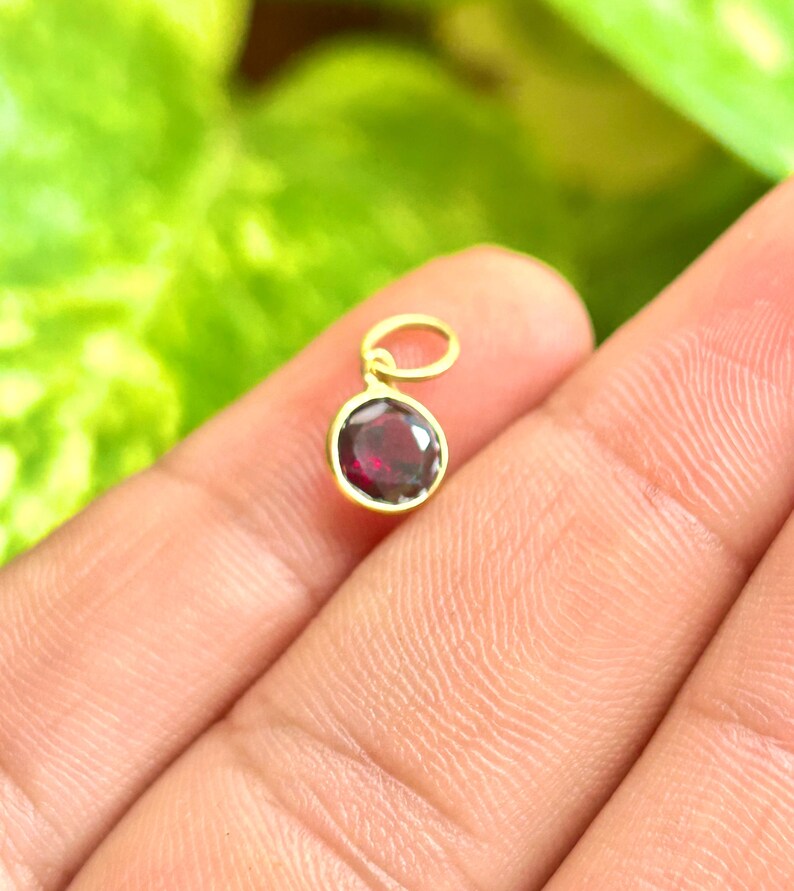 May include: A small, round pendant with a deep red gemstone set in a gold-colored bezel. The pendant has a loop at the top for attaching to a chain. The background is a blurred green.