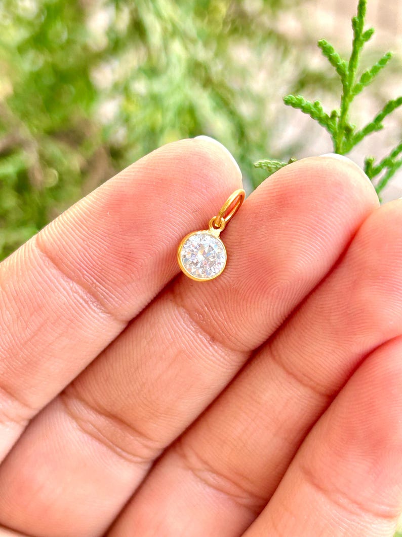 May include: A small, round gold pendant with a clear, sparkling gemstone. The pendant is held in a hand, with a blurred green background. The gold setting highlights the brilliance of the gemstone.