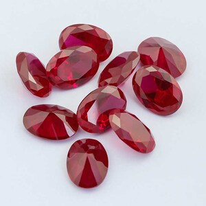 May include: A group of ten oval-shaped, faceted, red gemstones. The gemstones are arranged in a cluster on a white surface.