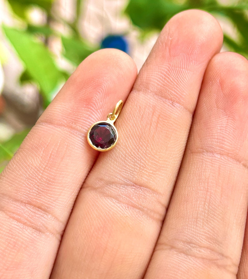 May include: A small, round pendant with a deep red gemstone set in a gold-colored bezel. The pendant has a loop at the top for a chain. The gemstone is a dark, rich color, and the gold setting is shiny.