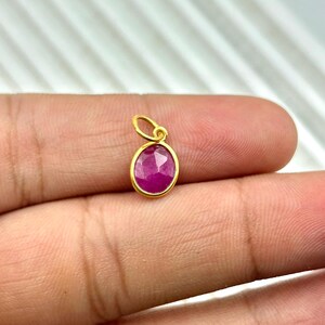 Puede incluir: Un pequeño colgante ovalado con una vibrante gema roja engastada en un marco de metal dorado. El colgante tiene un lazo para una cadena. La gema mide aproximadamente 1,3 cm de largo.