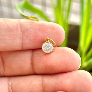 May include: A small, round gold pendant with a clear, sparkling gemstone. The pendant is held between fingers against a blurred green background. The gold setting has a small loop for a chain.