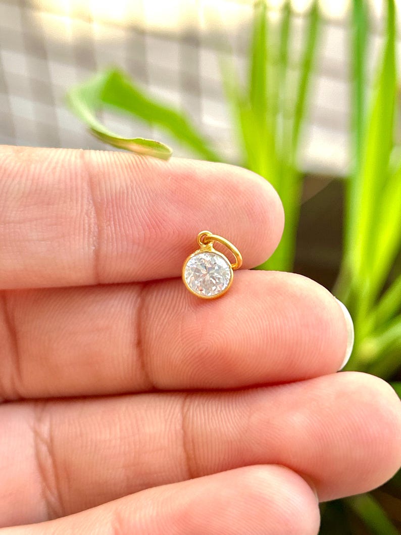 May include: A small, round gold pendant with a clear, sparkling gemstone. The pendant is held between fingers against a blurred green background. The gold setting has a small loop for a chain.