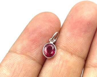 May include: A small, oval-shaped ruby pendant with a silver setting. The ruby is a deep red color and is set in a simple, elegant design. The pendant has a small loop at the top for attaching to a chain.