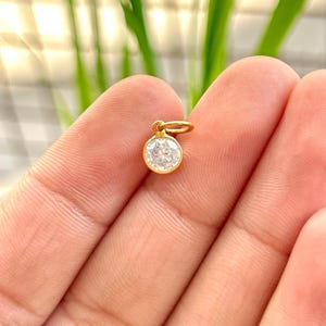 May include: A small, round, gold pendant with a clear, sparkling gemstone. The pendant is suspended from a small gold loop. The background is blurred, with green and white tones.
