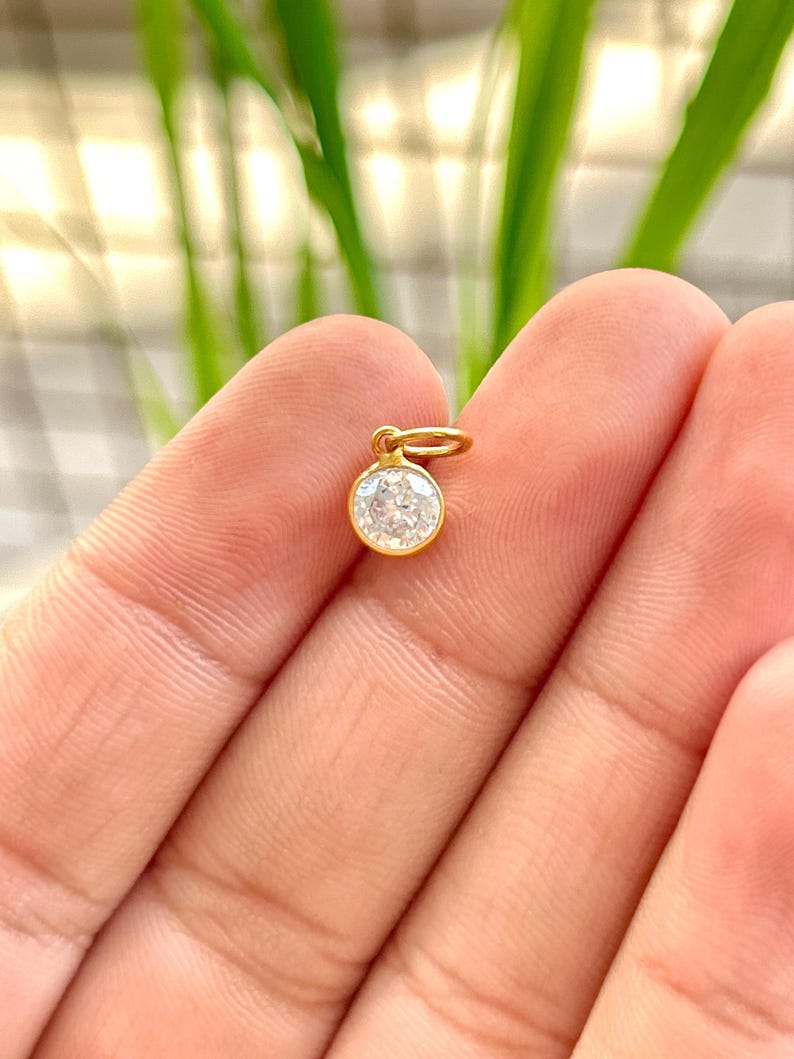 May include: A small, round, gold pendant with a clear, sparkling gemstone. The pendant is suspended from a small gold loop. The background is blurred, with green and white tones.