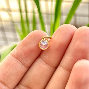 May include: A small, round, gold-colored pendant with a clear, faceted gemstone. The pendant is held between fingers against a blurred background of green foliage. The gemstone is circular and appears to be set in a gold bezel.