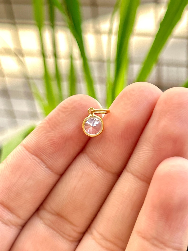 May include: A small, round, gold-colored pendant with a clear, faceted gemstone. The pendant is held between fingers against a blurred background of green foliage. The gemstone is circular and appears to be set in a gold bezel.
