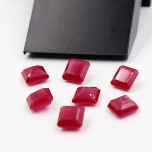 Może przedstawiać: Siedem czerwonych kwadratowych kamieni szlachetnych, prawdopodobnie rubinów, rozrzuconych na białym tle. Kamienie szlachetne są oszlifowane w kształcie kwadratu ze ściętymi krawędziami.