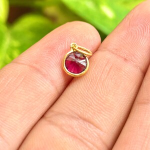 May include: A small, round gold pendant with a vibrant red gemstone. The gemstone is faceted, catching the light. The pendant has a simple, elegant design, perfect for a necklace or bracelet. The gold setting provides a warm contrast to the red stone.