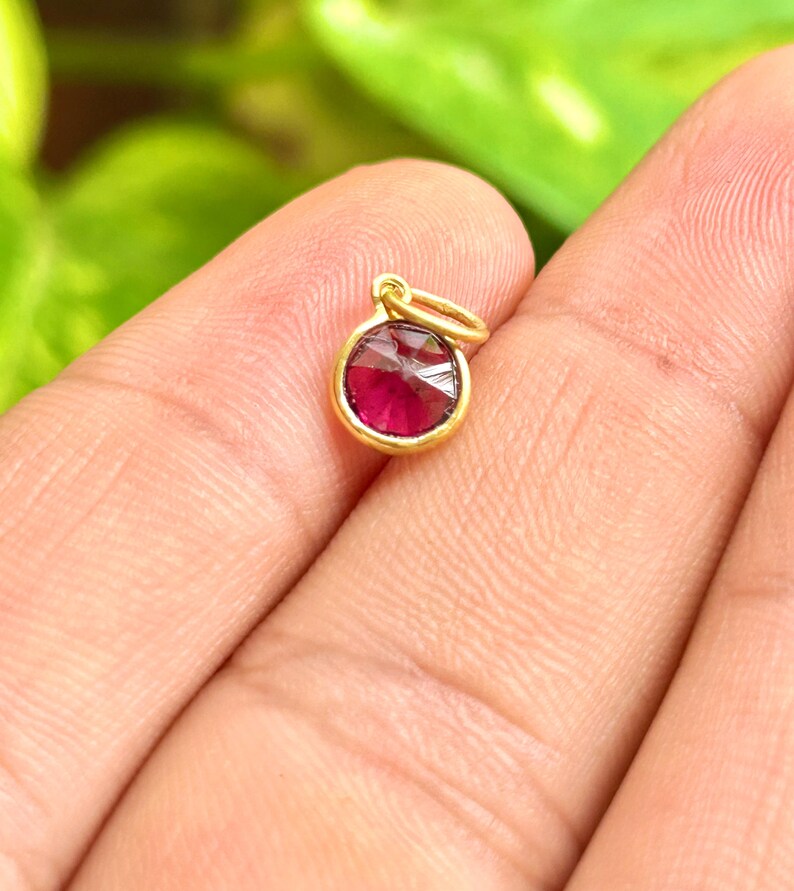 May include: A small, round gold pendant with a vibrant red gemstone. The gemstone is faceted, catching the light. The pendant has a simple, elegant design, perfect for a necklace or bracelet. The gold setting provides a warm contrast to the red stone.