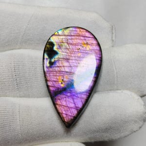 Puede incluir: Una piedra preciosa de labradorita en forma de lágrima pulida. La piedra muestra un vibrante juego de colores, con tonos de púrpura, rosa, azul y amarillo. La piedra preciosa está sobre un fondo blanco.