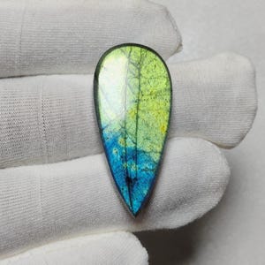 May include: A teardrop-shaped gemstone with a vibrant color gradient. The top half is a bright yellow-green, transitioning to a deep blue at the bottom. The stone has a polished surface with internal veining.