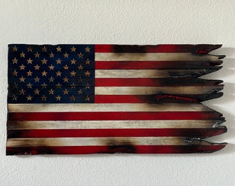 Puede incluir: Una bandera americana de madera envejecida con un diseño rústico de bordes quemados. La bandera presenta el campo azul tradicional con estrellas blancas y rayas rojas y blancas. La madera tiene una apariencia desgastada, lo que se suma a su estética vintage.