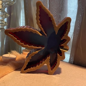 Może przedstawiać: Ozdobny przedmiot w kształcie liścia ze złotym konturem i ciemnoczerwonym i niebieskim wnętrzem. Liść spoczywa na jasnej powierzchni, z świecą i szklanym świecznikiem w tle.