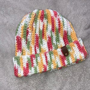 May include: A colorful crocheted beanie hat with a striped pattern in shades of pink, green, white, and yellow. The hat has a brown leather patch on the side.