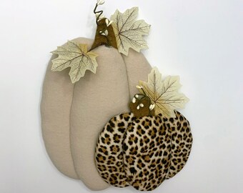 May include: Two fabric pumpkins for autumn decoration. The larger pumpkin is beige, the smaller one has a leopard print. Both have green stems and cream-coloured leaf accents. Ideal for seasonal home decor.