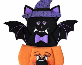 Puede incluir: Decoración de Halloween con un murciélago negro con un sombrero de bruja y una pajarita morados, posado sobre una calabaza naranja. El murciélago tiene alas forradas de morado y una cara sonriente. La calabaza tiene una cara sonriente.