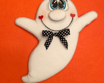 May include: A white ghost-shaped decoration with a smiling face, blue eyes, and a black and white polka dot bow tie. Red heart accents are on the cheeks. The background is a solid orange colour.