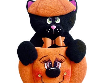 May include: A Halloween-themed plush decoration. It depicts a black cat wearing an orange pumpkin hat and bow tie, holding an orange pumpkin. The pumpkin has a smiling face with rosy cheeks and a black nose. The cat's hat is decorated with leaves and a small pumpkin.