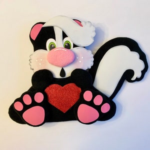 May include: A whimsical skunk-shaped decoration with a black body, white accents, and pink details. The skunk has a pink nose, ears, and paw pads, and a red glitter heart. The eyes are green. This charming piece is perfect for Valentine's Day.