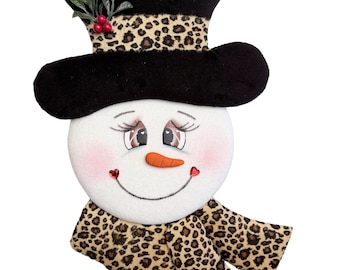May include: A snowman head decoration featuring a black top hat with a leopard print band, green holly leaves, and red berries. The white face has a carrot nose, pink cheeks, long eyelashes, and two small red heart-shaped accents at the smile.
