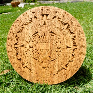 Könnte beinhalten: Eine runde Holzskulptur des Maya-Kalenders mit aufwendigen Details und einem zentralen Gesicht. Die natürliche Holzmaserung ist sichtbar. Die Skulptur liegt auf grünem Gras.
