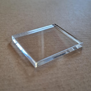Puede incluir: Una pieza cuadrada de acrílico transparente con bordes biselados. El material transparente refleja la luz, creando sutiles tonos de arcoíris. El objeto está posicionado sobre una superficie de tono neutro, mostrando sus líneas limpias y diseño minimalista.