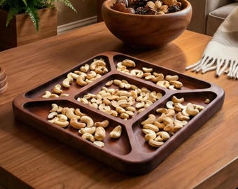 May include: A dark brown wooden serving tray with six compartments filled with cashews. A wooden bowl filled with pine cones and decorative items sits in the background. The tray is on a wooden table.