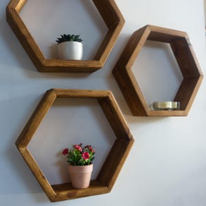 Peut inclure: Trois étagères hexagonales en bois fixées à un mur blanc. Chaque étagère est teintée d'une couleur marron chaud. L'une contient une petite plante succulente en pot, une autre une bougie et la troisième une petite plante en pot avec des fleurs roses.