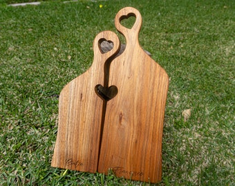 May include: Two wooden cutting boards shaped like people, standing side-by-side. The boards have heart-shaped cutouts and are engraved with the names "Antía" and "Francisco".