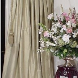 May include: A pair of pleated, light beige curtains hang in a window. The curtains are gathered at the top and have a tassel tieback. A vase of white and pink flowers sits on a dark wood table in the foreground.