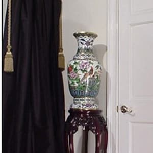 May include: A large, ornate, porcelain vase with a floral design sits on a dark wood pedestal stand. The vase is decorated with colorful flowers, birds, and geometric patterns. The stand has a carved design and four legs.