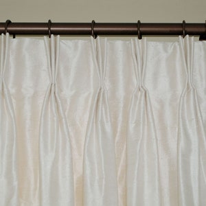 Puede incluir: Cortinas plisadas de color crema colgadas de una barra de cortina de bronce oscuro. Las cortinas tienen un brillo sutil y est&aacute;n recogidas a intervalos regulares, creando pliegues verticales. La barra tiene terminales decorativos.
