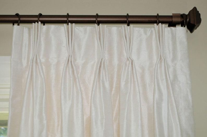 Puede incluir: Cortinas plisadas de color crema colgadas de una barra de cortina de bronce oscuro. Las cortinas tienen un brillo sutil y est&aacute;n recogidas a intervalos regulares, creando pliegues verticales. La barra tiene terminales decorativos.
