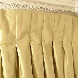 Puede incluir: Primer plano de una cortina plisada de color amarillo dorado. La tela tiene un ligero brillo y los pliegues crean pliegues verticales. La cortina cuelga de una moldura decorativa blanca y ornamentada.