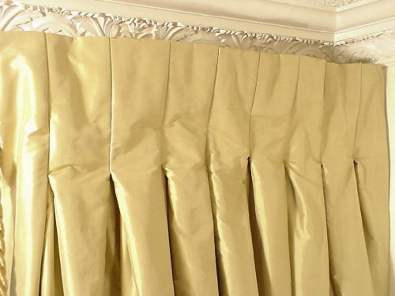 Puede incluir: Primer plano de una cortina plisada de color amarillo dorado. La tela tiene un ligero brillo y los pliegues crean pliegues verticales. La cortina cuelga de una moldura decorativa blanca y ornamentada.