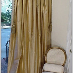 May include: Gold pleated curtains with tassels hanging from the top, covering a window with a view of a street. A chair with a white cushion sits in the corner of the room.