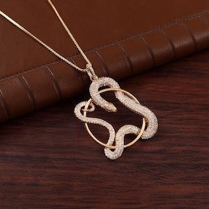 May include: A gold-toned necklace featuring a pendant shaped like a snake coiled around a circular hoop. The snake is encrusted with numerous small, clear, sparkling stones that cover its entire surface, suspended from a delicate gold-toned chain.
