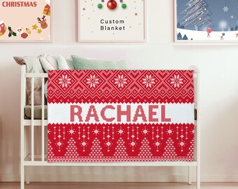 May include: A red and white Christmas-themed blanket with a white stripe that says "RACHAEL" in red letters. The blanket is on a white crib.