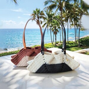 May include: Two handmade crochet tote bags with leather handles. One bag is a mix of white, black, and cream colors, while the other is a mix of brown and cream colors. The bags are sitting on a white surface with a beach and palm trees in the background.
